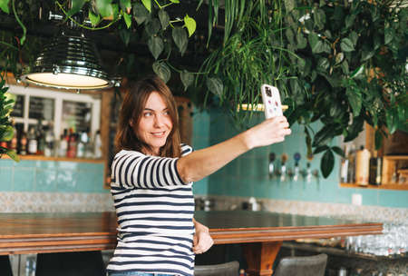Young smiling brunette woman in casual clothes using social media on mobile phone sitting at green...