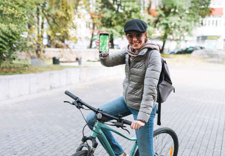 Young smiling brunette woman in casual clothes using social media on mobile phone sitting at green...