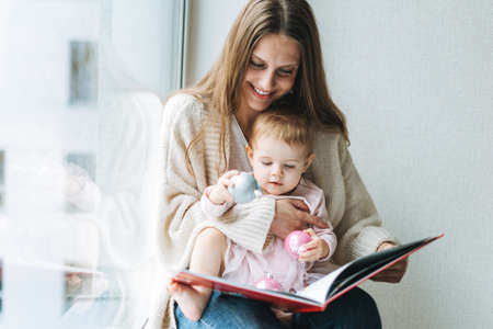 Young woman mom with long hair in cozy knitted cardigan with baby girl on hands near window at homeの写真素材