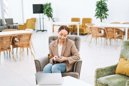 Adult smiling brunette woman forty years in headphones doing notes in daily book with opened laptop....