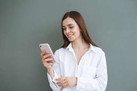 Portrait of beautiful smiling woman student with dark long hair in white shirt using mobile phone in hand on gray background, generation Zの写真素材