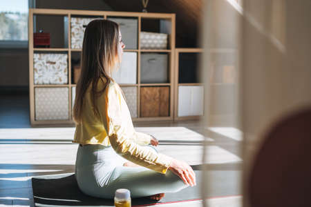 Young beautiful woman forty year with blonde long curly hair in cozy knitted gray sweater with mobile phone in hands sitting on couch in bright interior at homeの写真素材