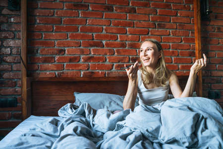 Adult woman forty years old with blonde long hair in pajamas using mobile phone sitting on bed in loft room at homeの写真素材