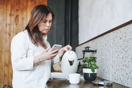 Adult middle aged brunette smiling woman fifty years in white shirt with cup of morning coffee in kitchen at homeの写真素材