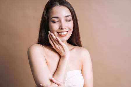 Beauty nude portrait of happy young beautiful asian woman with healthy dark long hair in top bando on beige background isolatedの写真素材