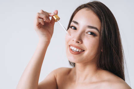 Beauty portrait of happy smiling asian woman with dark long hair with pipette with organic oil on clean fresh skin face and hands on white background isolatedの写真素材