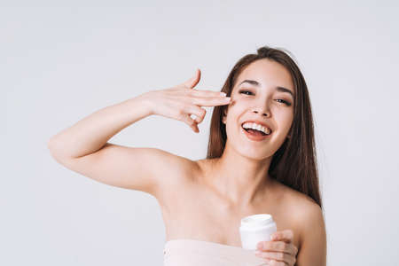 Beauty portrait of happy smiling asian woman with dark long hair with pipette with organic oil on clean fresh skin face and hands on white background isolatedの写真素材