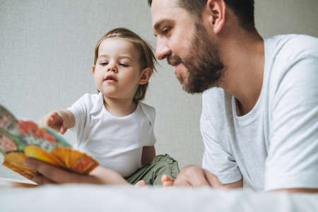 Happy father young man feeds baby girl little daughter in kitchen at homeの写真素材