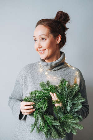 Young woman in warm gray knitted dress making diy fir christmas wreath in hand on the table at homeの写真素材