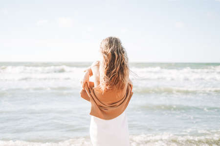 Young beautiful woman with long hair in beige sweater enjoying life on sea beach at sunset