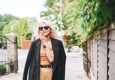 Adult elegant blonde woman in black jacket and sunglasses on summer city street