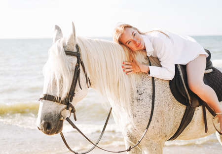 Young red haired woman in white shirt riding white horse on seascape backgroundの写真素材