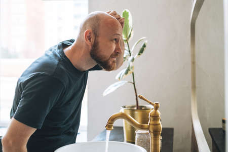 Handsome bald man looking at mirror and touching face in bathroom
