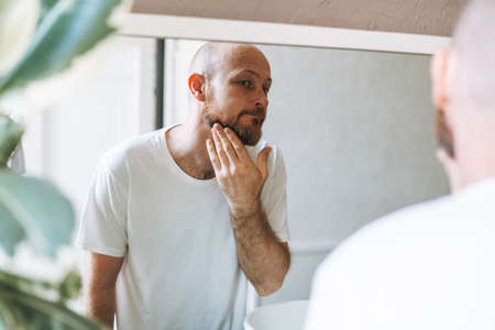 Handsome bald man looking at mirror and touching face in bathroomの写真素材