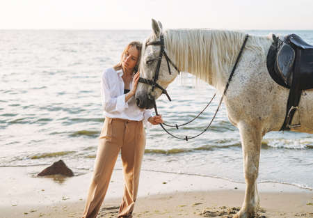 Young long hair woman in white shirt riding white horse on seascape background