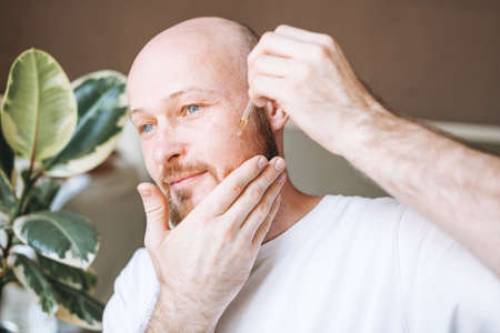 Handsome young bearded man trimming his beard with machine in bathroom at homeの写真素材