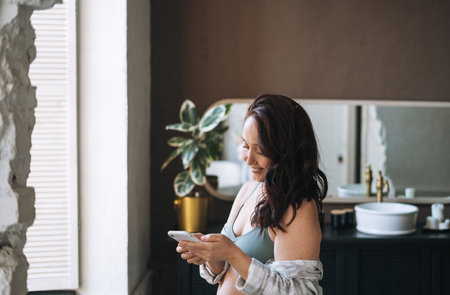 Self loving plus size middle aged woman in underwear using mobile phone in bathroom at home