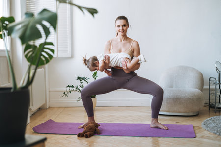 Young woman fit mom with baby girl in her arms doing fitness on mat at homeの写真素材