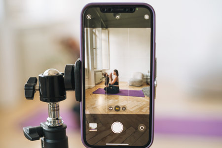 Young woman with little daughter fitness blogger doing exercises and recording video on mobile phone in studioの写真素材