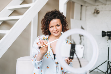 Young brunette woman blogger in home clothes doing makeup with mobile phone and ring lamp online in studioの写真素材