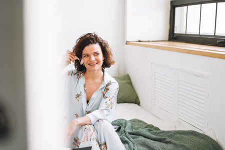 Young brunette woman in blue pajamas doing face massage with stone roller sitting on bed at homeの写真素材