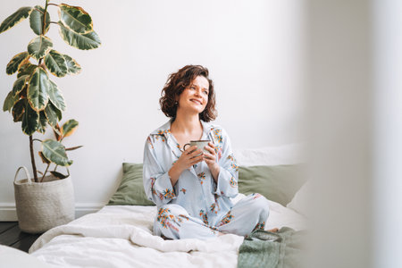 Young smiling woman with brunette curly hair in blue pajamas with cup of tea in hands sitting on bed at homeの写真素材