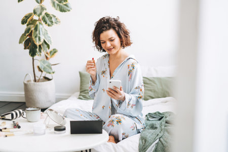 Young brunette woman in blue pajamas applies makeup with mobile phone sitting on bed at homeの写真素材
