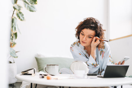 Young brunette woman in blue pajamas applies makeup with mobile phone sitting on bed at homeの写真素材