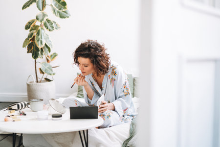 Young brunette woman in blue pajamas applies makeup in front of mirror sitting on bed at homeの写真素材