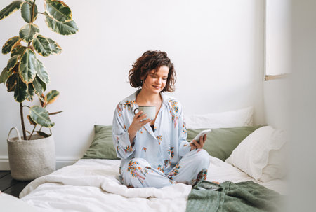 Young brunette woman with curly hair in pajamas using mobile on bed at homeの写真素材