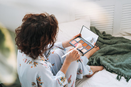 Young brunette woman in blue pajamas applies makeup with face sculpting palette sitting on bed at homeの写真素材