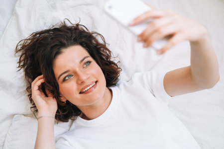 Young smiling brunette woman in home clothes using mobile on bed at home, top viewの写真素材