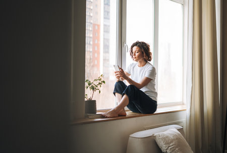 Young brunette woman in casual clothes using mobile phone sitting on window sill at homeの写真素材