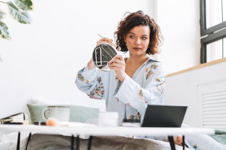 Young brunette woman in blue pajamas applies makeup in front of mirror sitting on bed at homeの写真素材