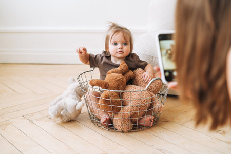 Cute baby girl sitting in basket with soft toys and young mother taking photo on mobile phone at homeの写真素材