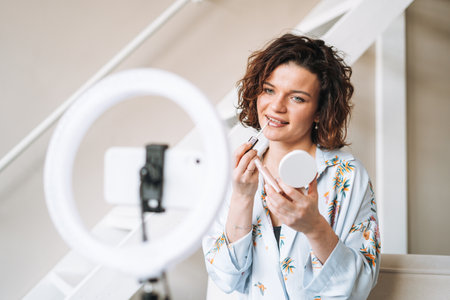 Young brunette woman blogger in home clothes doing makeup with mobile phone and ring lamp online in studioの写真素材