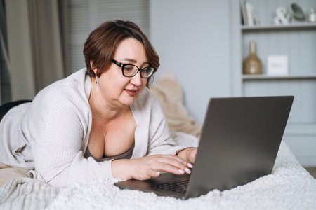 Smiling middle aged plus size woman lying on bed, working on laptop from homeの写真素材