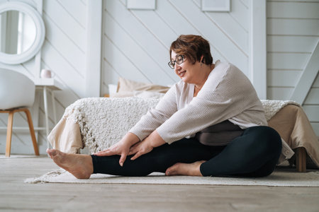 Middle aged plus size smiling brunette woman in home wear doing stretching in bedroomの写真素材