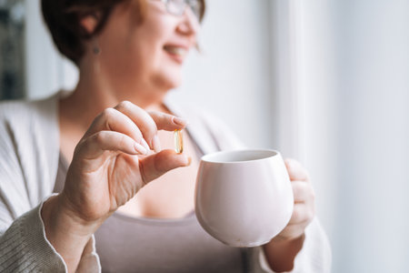 Close up photo of middle aged woman holding omega 3 capsule and mug of water in hands near window at homeの写真素材