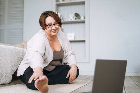 Adult charming brunette woman plus size body positive practice yoga with laptop at homeの写真素材