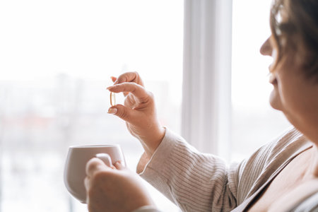 Close up photo of middle aged woman holding omega 3 capsule and mug of water in hands near window at homeの写真素材
