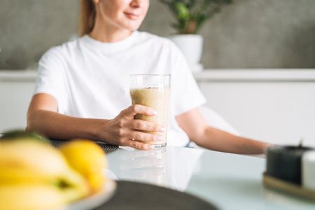 Young slim woman in white t-shirt and blue jeans drinking fruit smoothie healthy food in kitchen at homeの写真素材