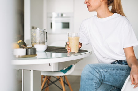 Young slim woman in white t-shirt and blue jeans drinking fruit smoothie healthy food in kitchen at homeの写真素材
