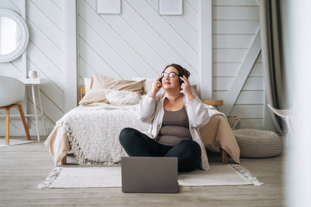 Middle aged plus size smiling brunette woman in home wear listening music in headphones using laptop in bedroomの写真素材