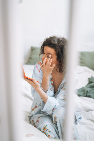 Young brunette woman in blue pajamas applies makeup with eye shadow palette in hands at homeの写真素材