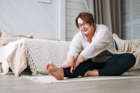 Middle aged plus size smiling brunette woman in home wear doing stretching in bedroomの写真素材