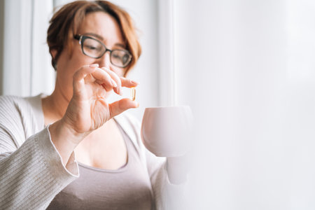 Close up photo of middle aged woman holding omega 3 capsule and mug of water in hands near window at homeの写真素材