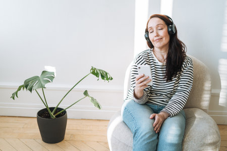 Smiling brunette woman with closed eyes using mobile phone, listen music in the headphones in chair at homeの写真素材
