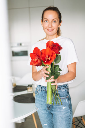 Portrait of young woman in white t-shirt with bouquet of red flowers in hands near window in bright kitchen at homeの写真素材