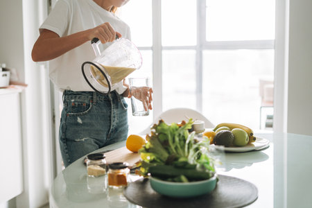 Young slim woman in white t-shirt and blue jeans pouring fruit smoothie healthy food in kitchen at homeの写真素材